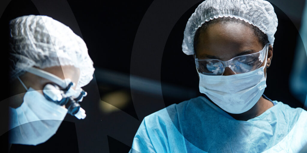 Two surgeons at work together, wearing surgical masks, hair nets, and blue scrubs.