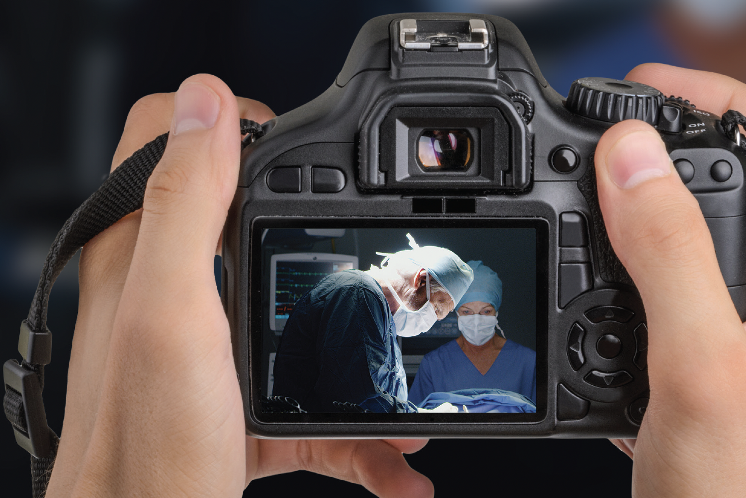 Hands holding a digital camera, the viewfinder on which shows two surgeons working together.